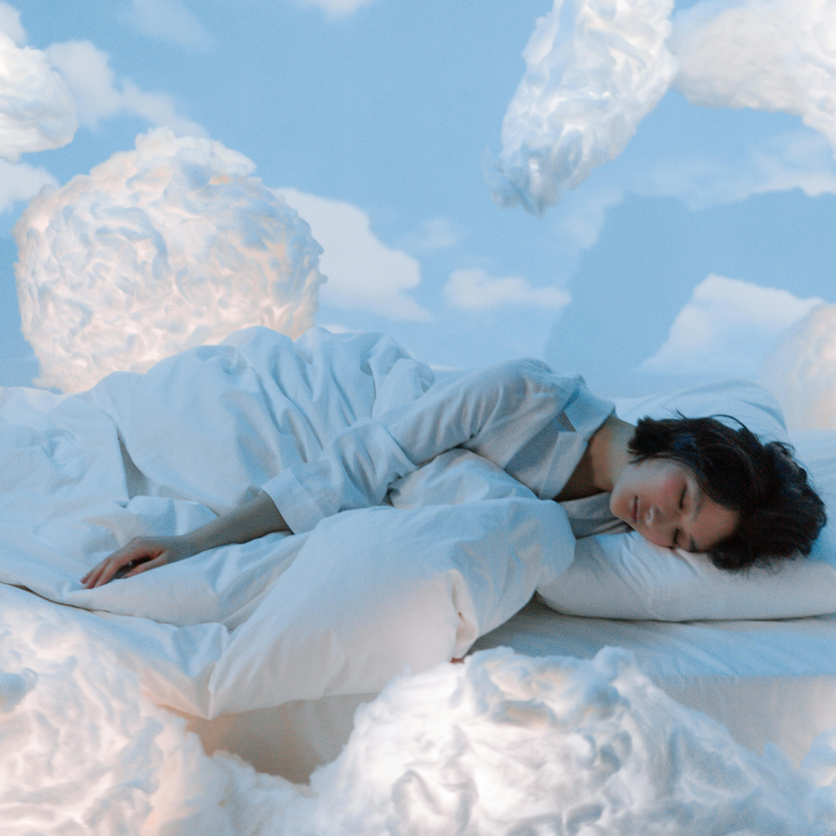 Kobieta śpi w białej pościeli, otoczona jest chmurami na niebieskim tle