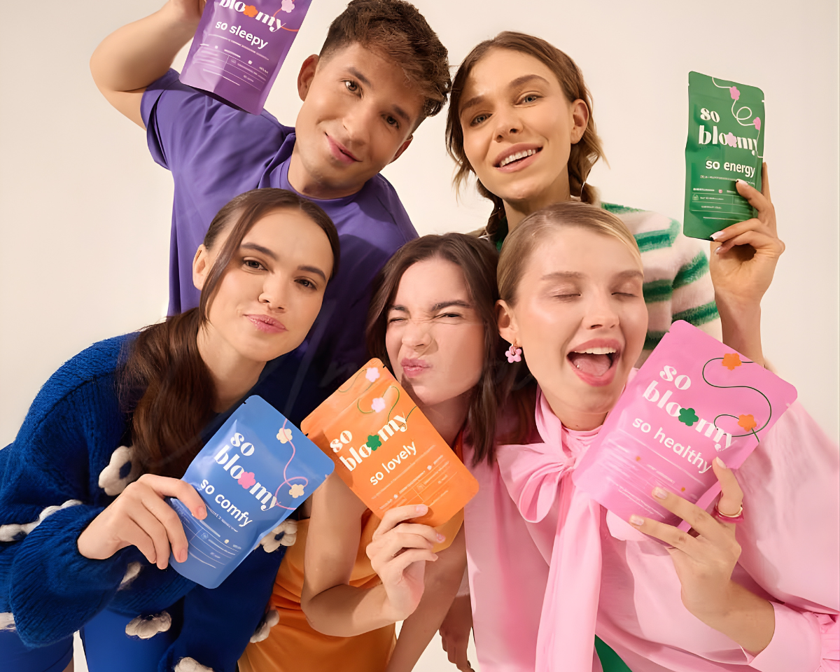 Pięć młodych osób uśmiecha się do zdjęcia, każda z tych osób trzyma w dłoni po jednym opakowaniu suplementów w żelkach so bloomy.