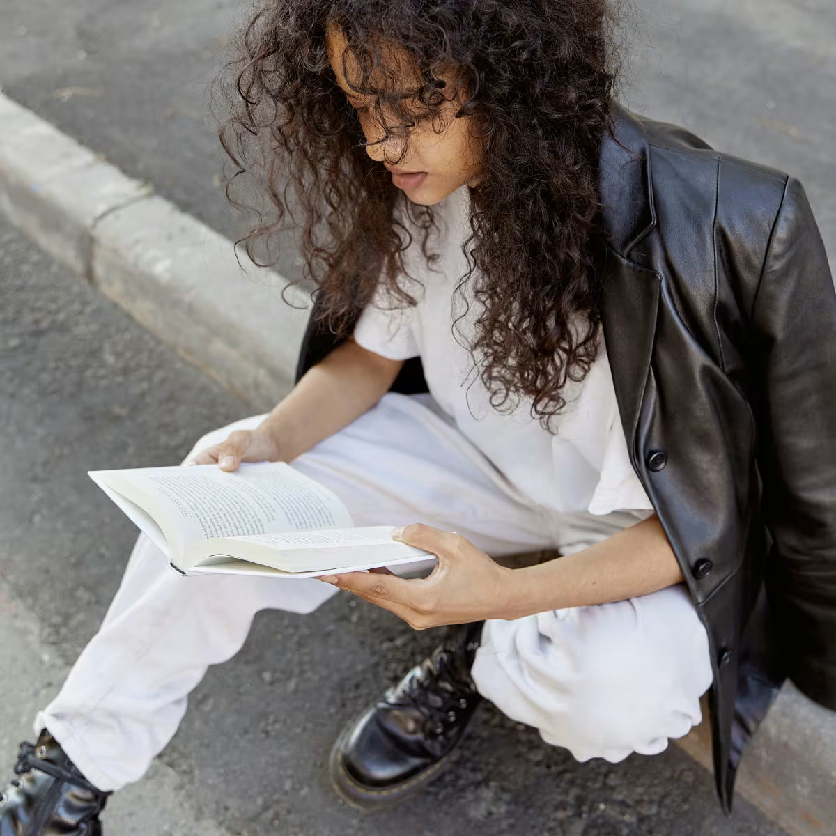Stylowo ubrana dziewczyna siedzi na krawężniku i czyta książkę.