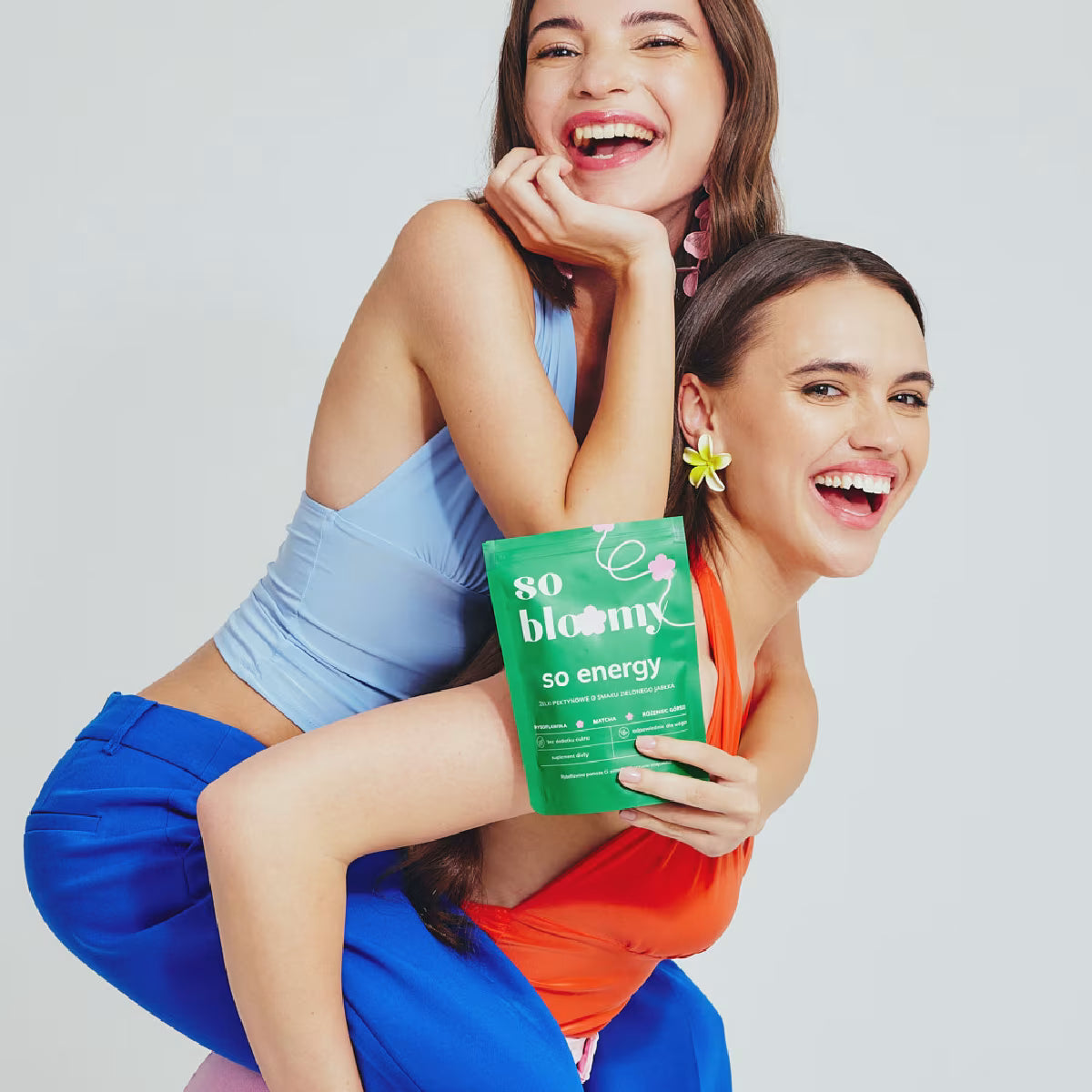 Dwie ciemnowłose dziewczyny z promienistymi cerami pozują na jasnym tle. Jedna dziewczyna trzyma drugą na plecach uśmiechając się szeroko. Jedna z dziewczyn trzyma w dłoni opakowanie wegańskich żelków bez cukru od marki so bloomy.
