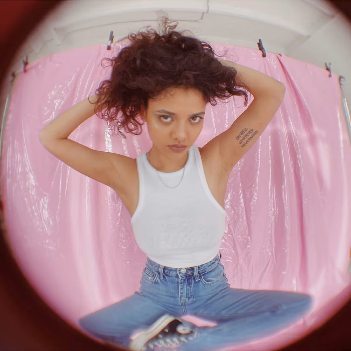 Dziewczyna ubrana w jeansy i trampki trzyma się za włosy stojąc na różowym tle. Zdjęcie zrobione z efektem rybiego oka.