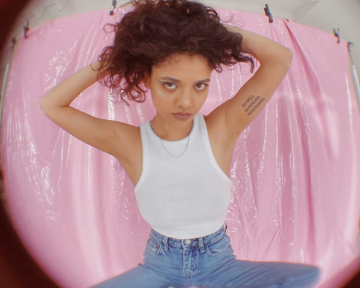 Dziewczyna ubrana w jeansy i trampki trzyma się za włosy stojąc na różowym tle. Zdjęcie zrobione z efektem rybiego oka.