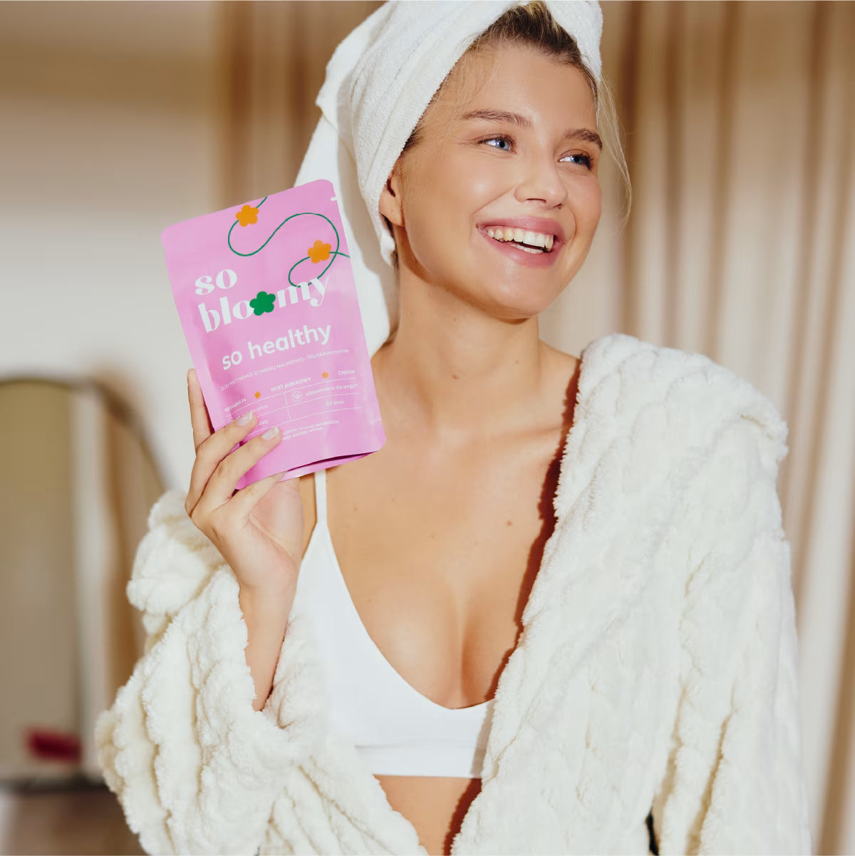 Uśmiechnięta dziewczyna o zdrowej i promiennej cerze, ubrana w szlafrok i ręcznik na włosach, uśmiecha się szeroko, a w dłoni trzyma wegańskie żelki so bloomy.