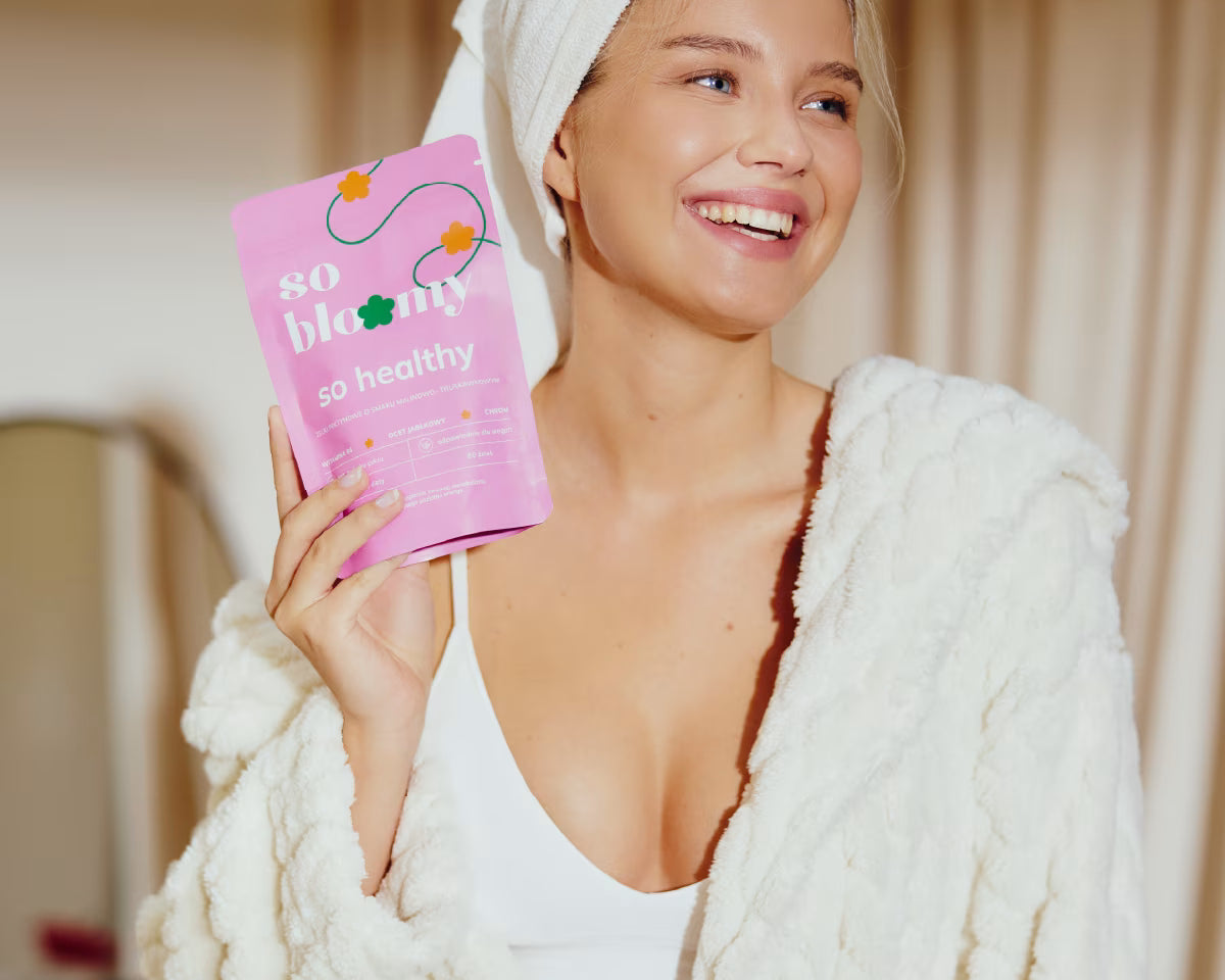 Uśmiechnięta dziewczyna o zdrowej i promiennej cerze, ubrana w szlafrok i ręcznik na włosach, uśmiecha się szeroko, a w dłoni trzyma wegańskie żelki so bloomy.