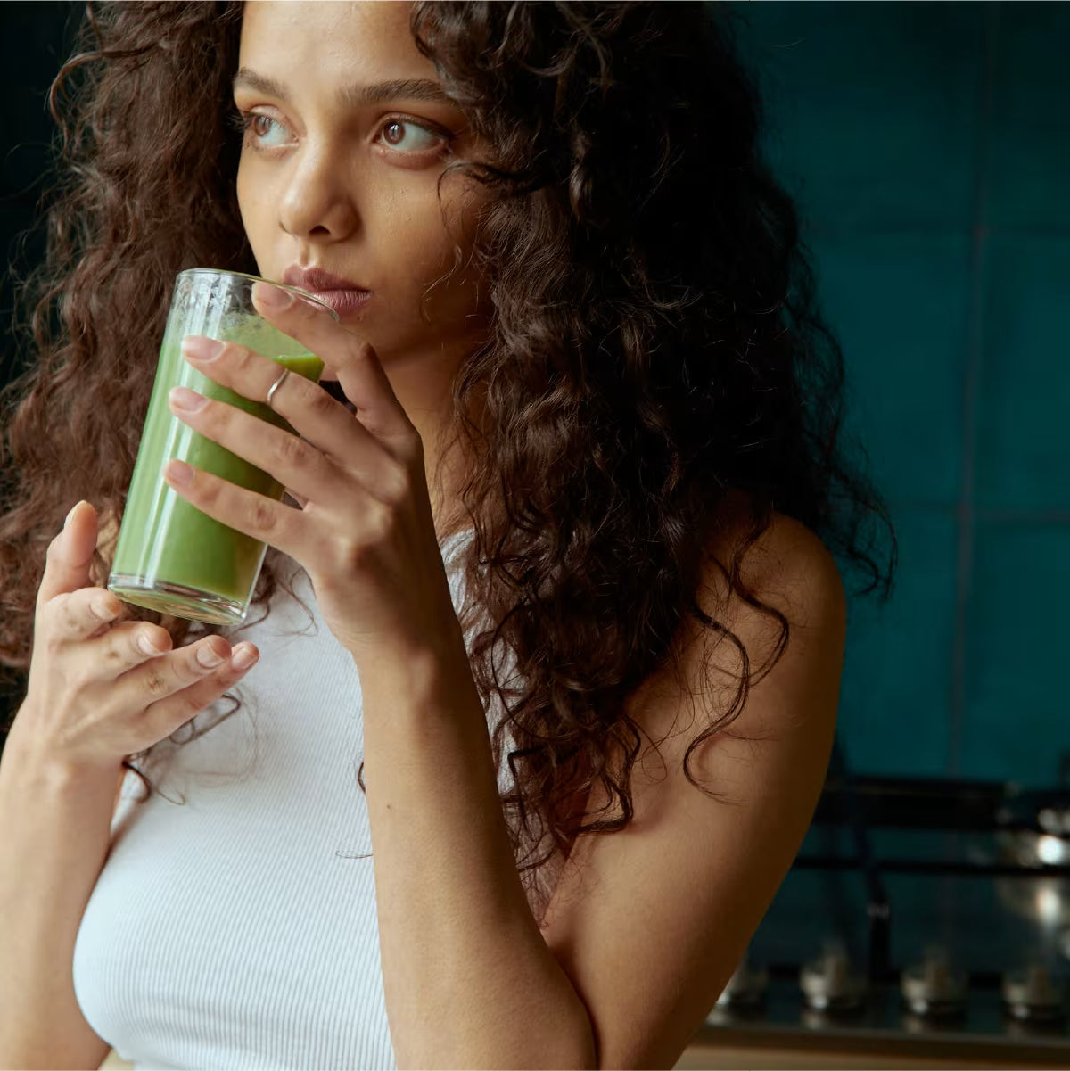 Dziewczyna o ciemniejszej karnacji, ubrana na biało, pije zielony koktajl i patrzy w dal.