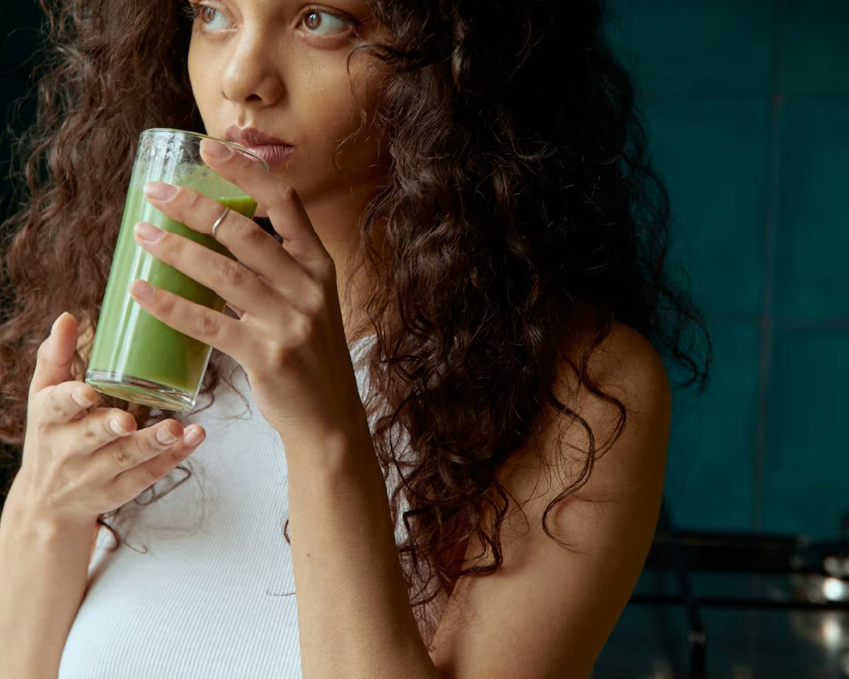 Dziewczyna o ciemniejszej karnacji, ubrana na biało, pije zielony koktajl i patrzy w dal.
