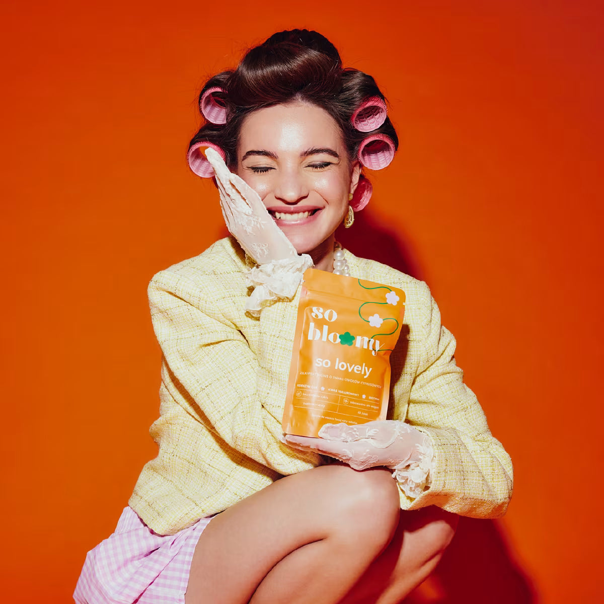 Uśmiechnięta kobieta z wałkami we włosach kuca na pomarańczowym tle. Jedną dłoń trzyma na promiennej twarzy, a w drugiej dłoni trzyma opakowanie żelek bez cukru so bloomy. 