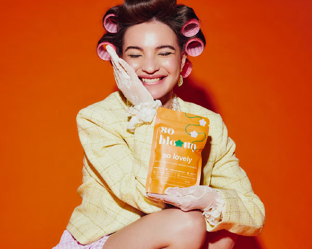 Uśmiechnięta kobieta z wałkami we włosach kuca na pomarańczowym tle. Jedną dłoń trzyma na promiennej twarzy, a w drugiej dłoni trzyma opakowanie żelek bez cukru so bloomy. 