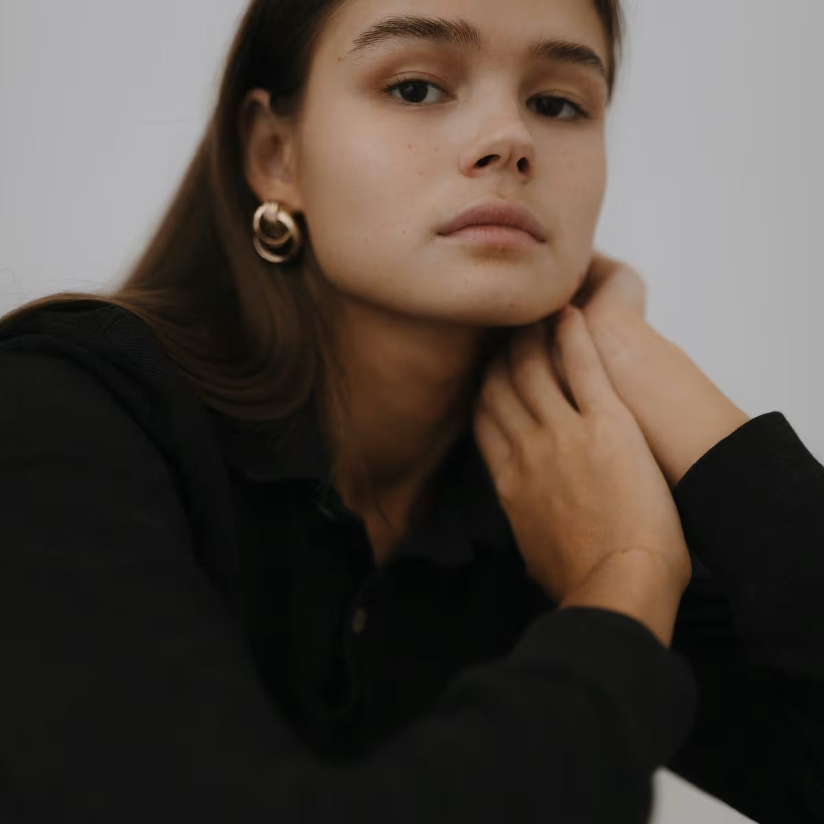 Dziewczyna z gładką cerą i długimi włosami patrzy się w obiektyw trzymając dłonie pod twarzą. 