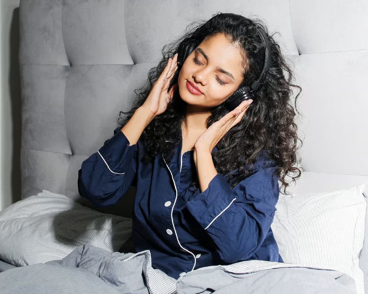 Ciemnowłosa dziewczyna siedzi na łóżku w piżamie i słucha na słuchawkach muzyki z zamkniętymi oczami.