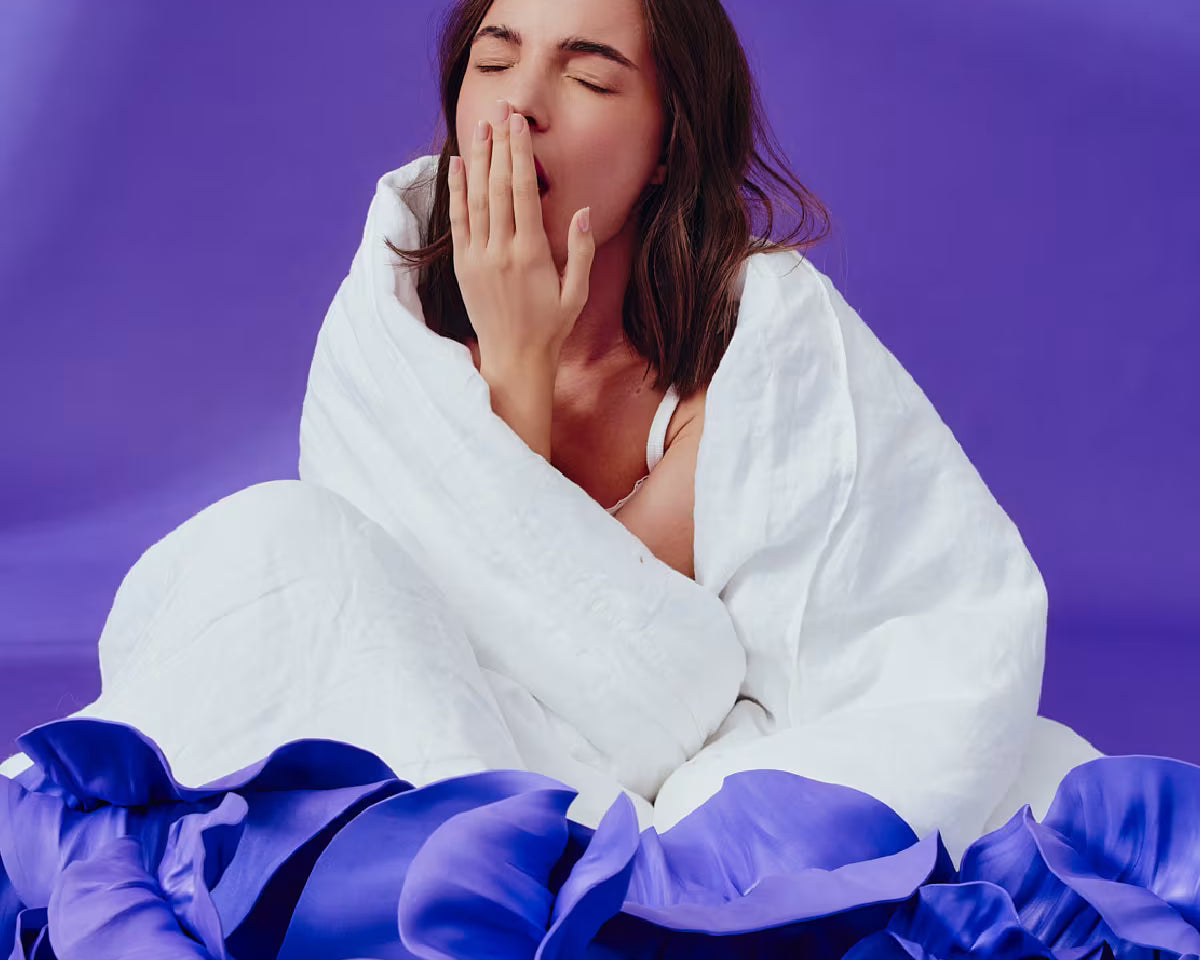 Dziewczyna z brązowymi włosami owinięta białą pościelą ziewa zasłaniając usta dłonią. Otoczona jest fioletowym tłem i fioletowymi kwiatami.