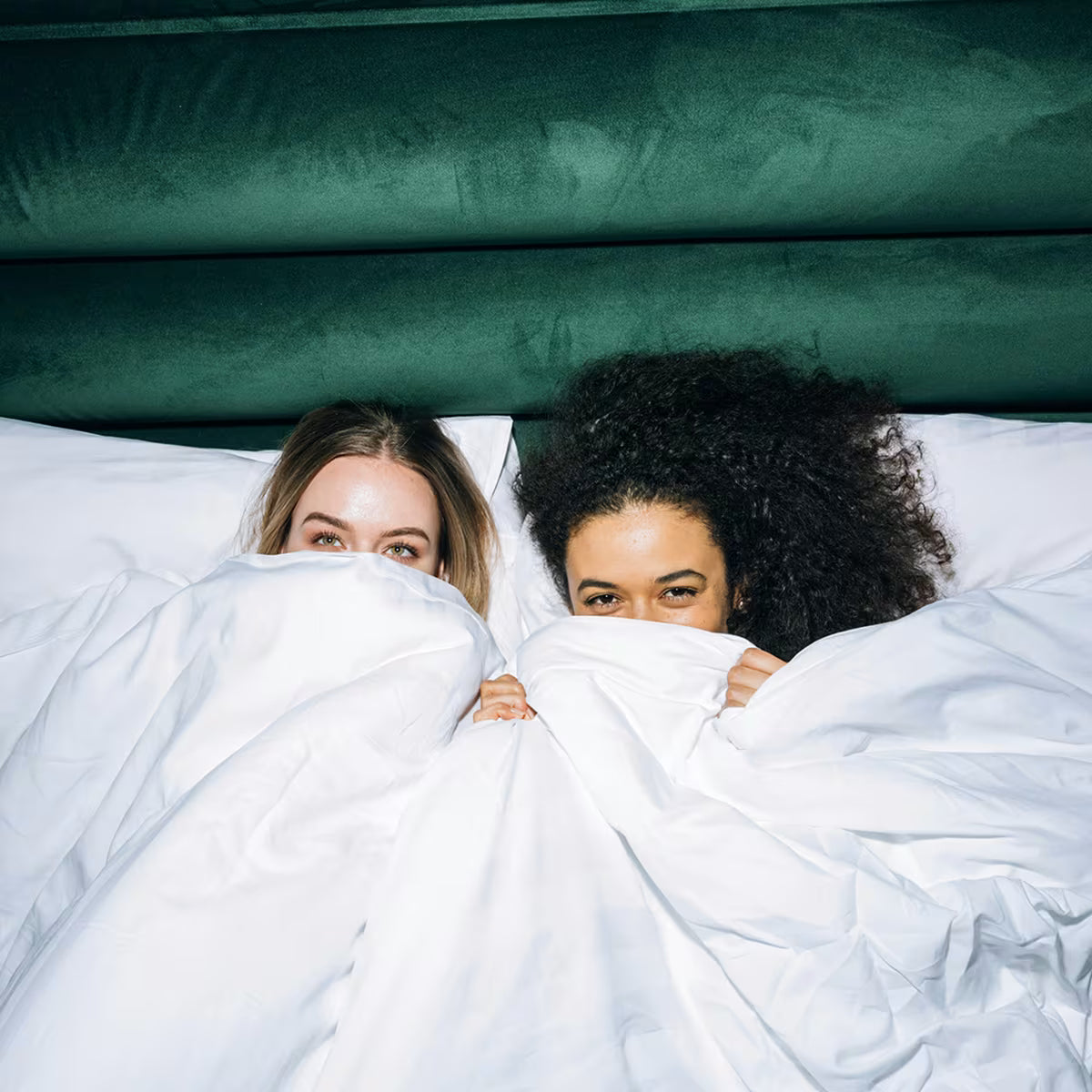 Dwie kobiety leżące obok siebie w łóżku, częściowo zakrywając twarze białą kołdrą