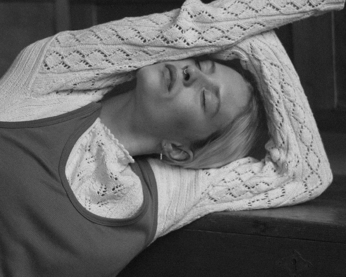 Kobieta opierająca się o stół z zamkniętymi oczami i rękami uniesionymi nad głową, wyglądająca na zmęczoną i zamyśloną.