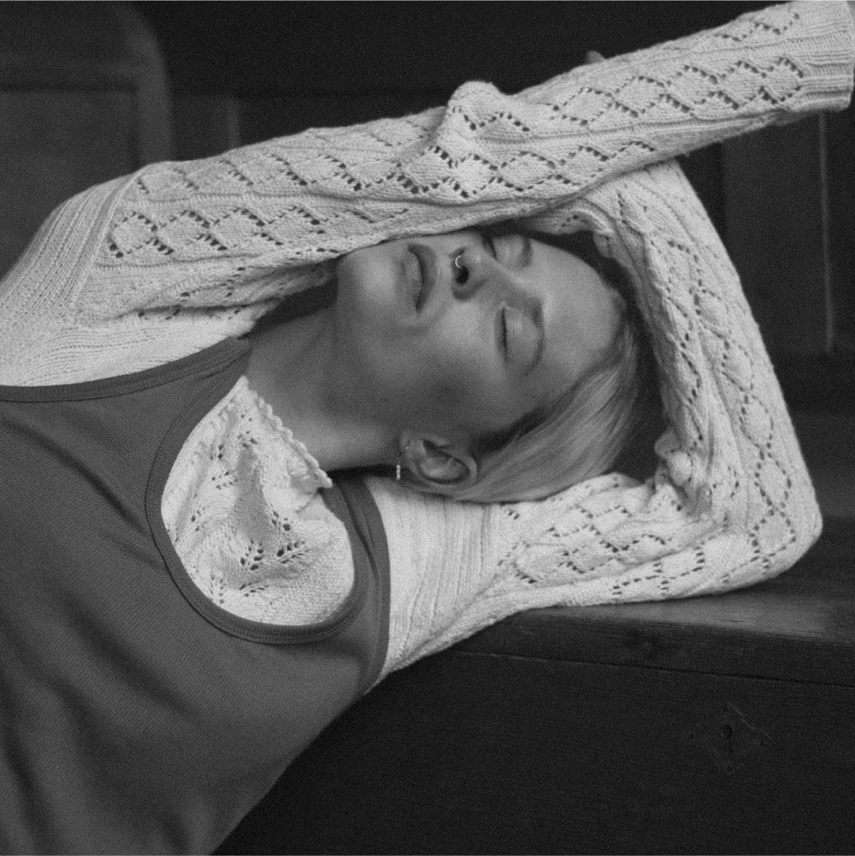 Kobieta opierająca się o stół z zamkniętymi oczami i rękami uniesionymi nad głową, wyglądająca na zmęczoną i zamyśloną.