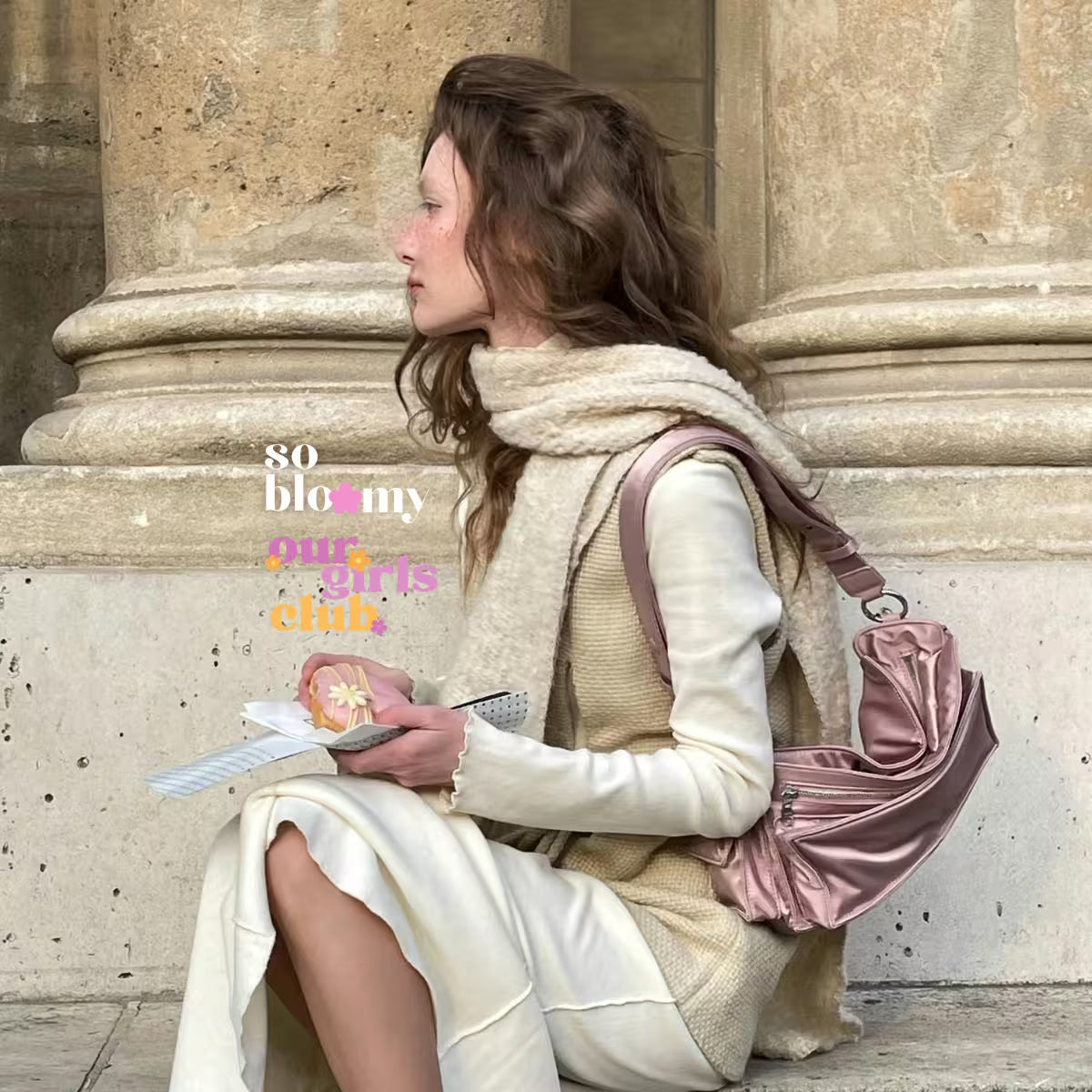 Elegancka kobieta ubrana w jasnych kolorach siedzi na schodach przed budynkiem. W dłoniach trzyma ciastko z kwiatkiem oraz notes. Na zdjęciu widoczne logo Our Girls Club oraz so bloomy.