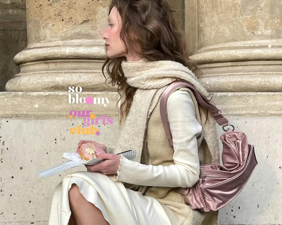 Elegancka kobieta ubrana w jasnych kolorach siedzi na schodach przed budynkiem. W dłoniach trzyma ciastko z kwiatkiem oraz notes. Na zdjęciu widoczne logo Our Girls Club oraz so bloomy.