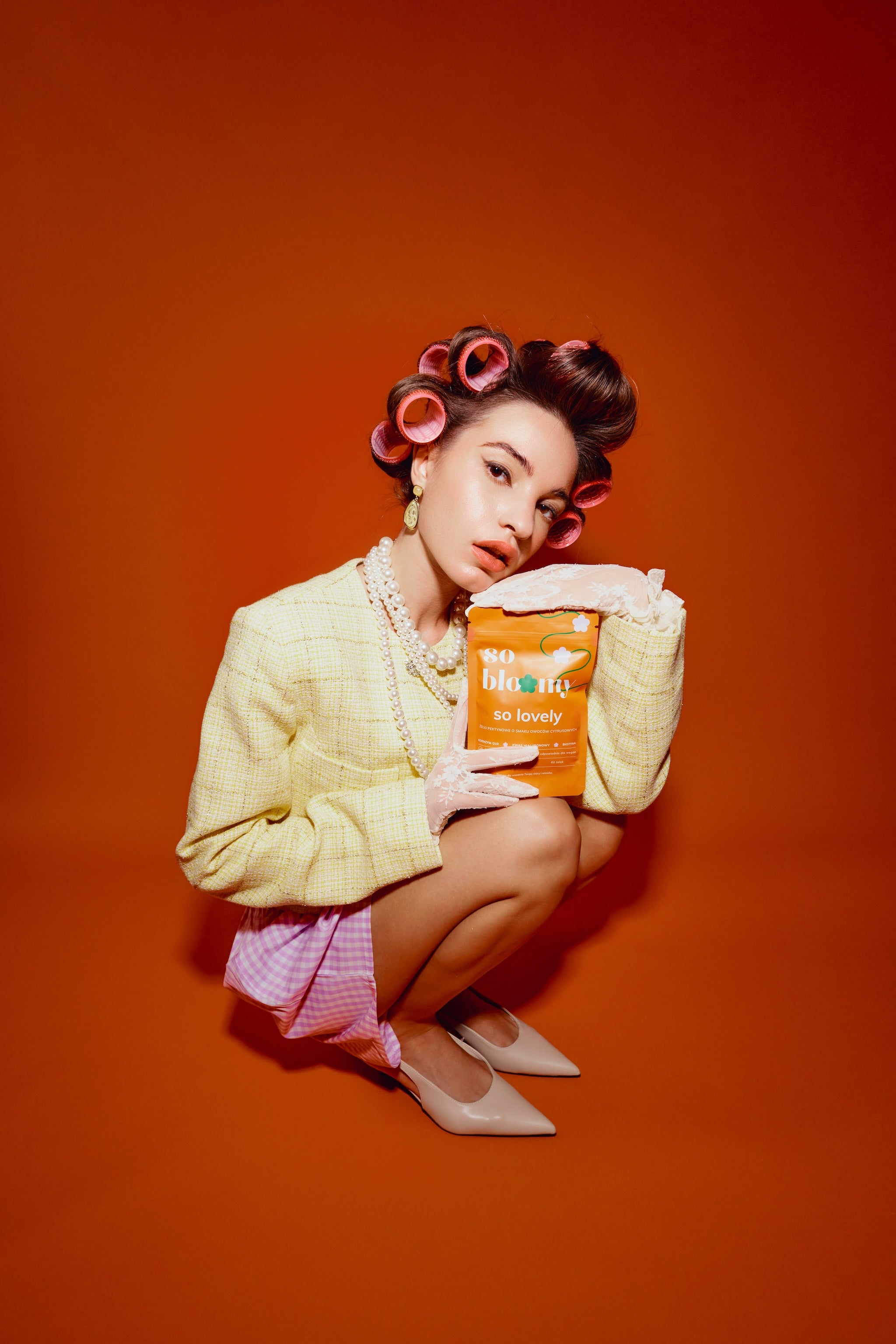Elegancko ubrana dziewczyna w wałkach we włosach kuca na pomarańczowym tle, w dłoniach trzyma opakowanie żelek so lovely